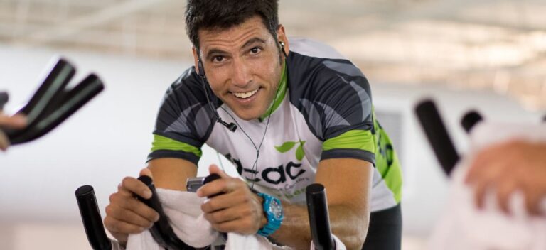Person leaning forward on a stationary bike, wearing an ACAC-branded cycling jersey and earphones, with a towel draped over the handlebars in an indoor fitness setting.