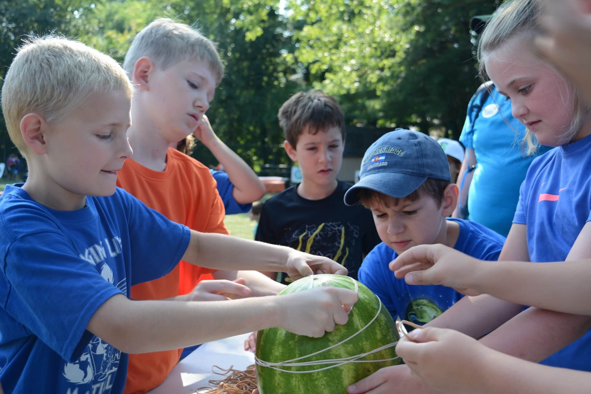 Children are gathered around a table outdoors, working together to wrap rubber bands around a large watermelon. The activity appears to be a group experiment or challenge, with several hands actively placing bands on the fruit. Trees and greenery are visible in the background, suggesting a camp or outdoor event setting.